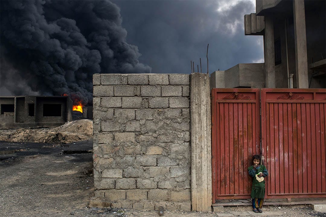 2016年11月10日，伊拉克盖亚拉，起火的油井附近，一个女孩站在家门外吃面包。