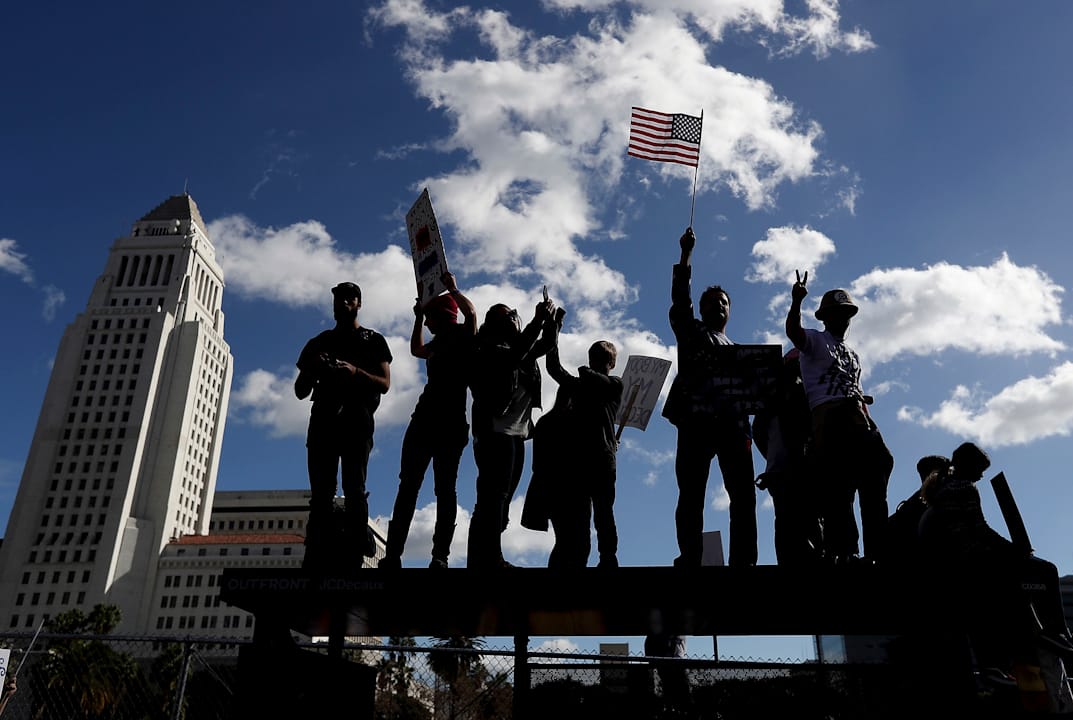 在美国加洲洛杉矶，有抗议者爬上巴士站上高举标语牌。