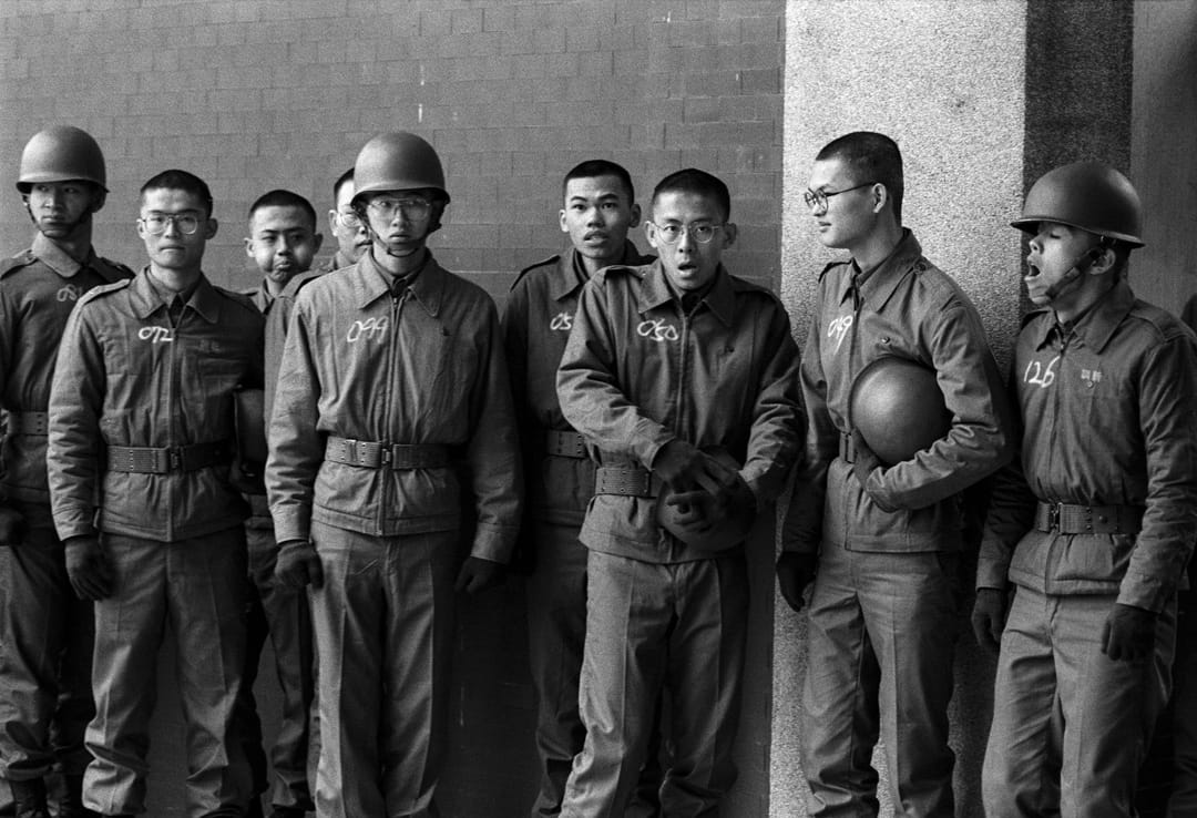 一群剛報到幾天,排隊等待拍攝大頭照的大專集訓學生,衣服上用粉筆寫着學號,個個表情是菜鳥,面對軍中環境的茫然。